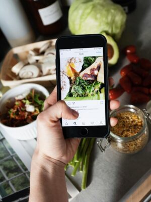 From above of crop person touching cellphone screen while searching for image of healthy recipe and standing in kitchen in front of vegetables