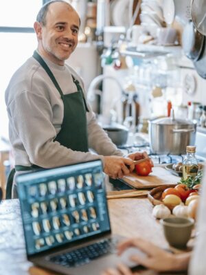 Smiling man cutting tomato against crop partner with laptop